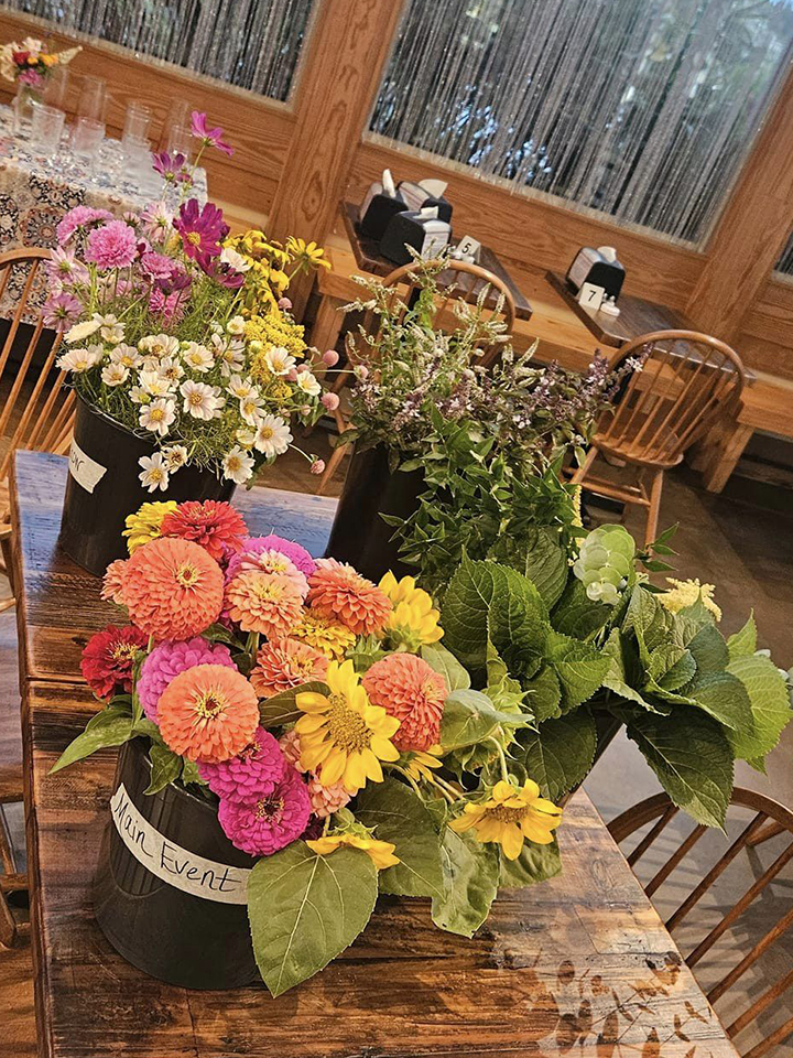 a table covered in large buckets of flowers and greenery with a table of vases in the background