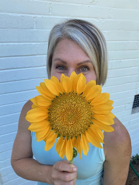 a woman holding a sunflower in front of her face with smiling eyes peeking over the top
